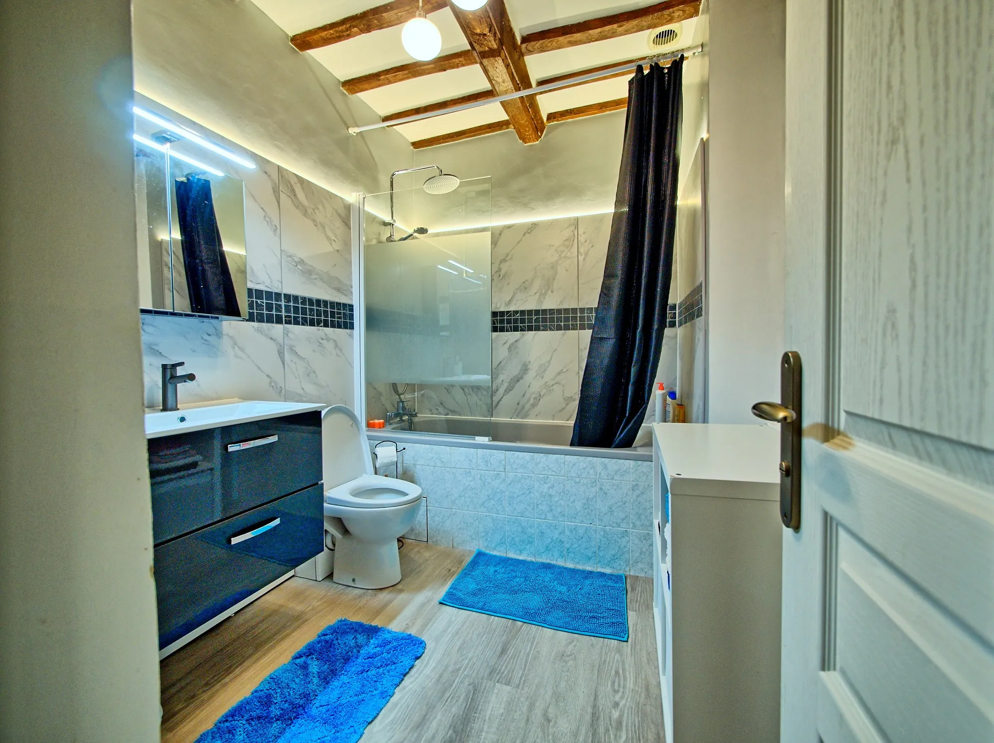 Salle de bain refaite : baignoire avec douche de pluie, faïence marbrée, poutres apparentes